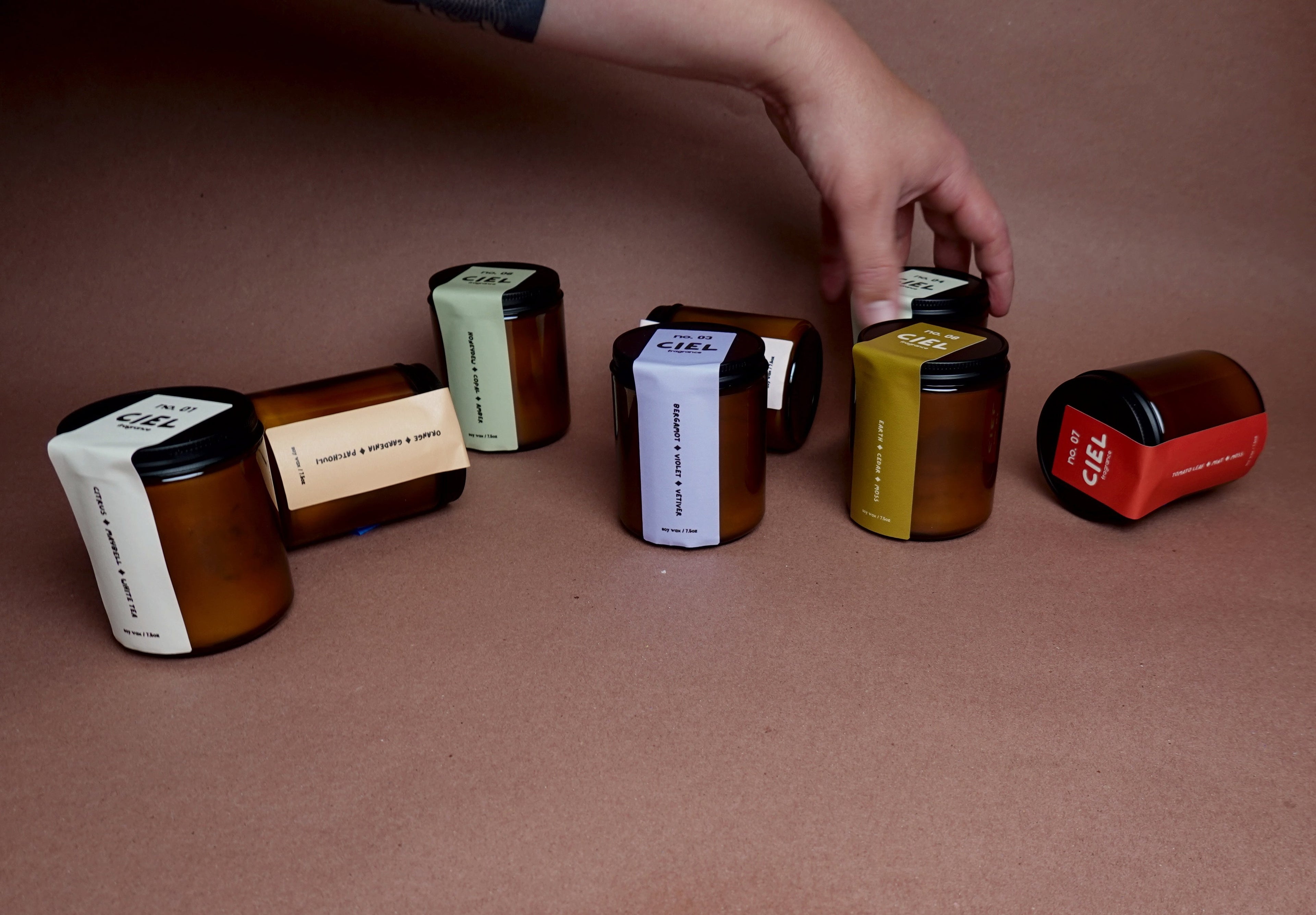 Hand reaching towards a row of amber candle jars with labels on a brown surface